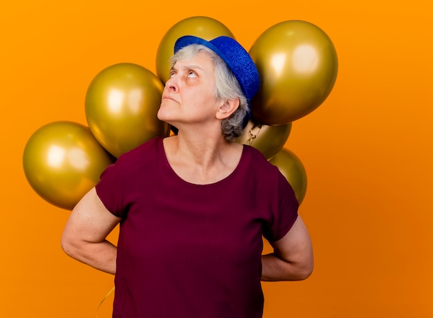 Annoyed elderly woman wearing party hat holds helium balloons behind looking up on orange