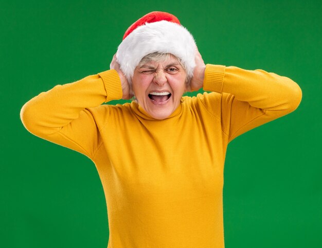 Annoyed elderly woman with santa hat covering ears with hands isolated on purple wall with copy space