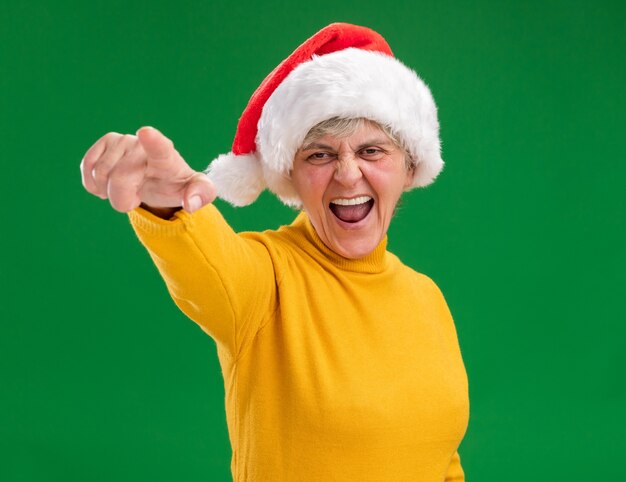 annoyed elderly woman with santa hat looking and pointing at camera isolated on purple background with copy space