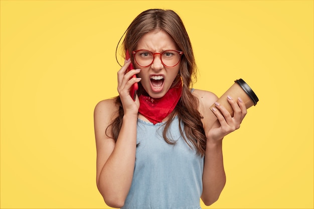 Annoyed European woman yells with negative feelings while has telephone conversation, argues  carries coffee in disposable cup