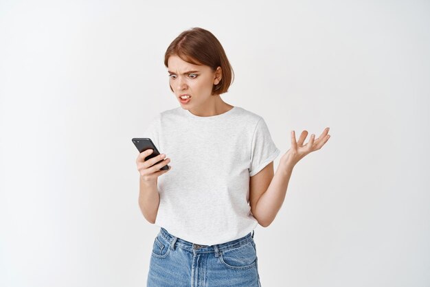 Annoyed girl look at smartphone screen and complaining staring angry at display with raised hand standing against white background