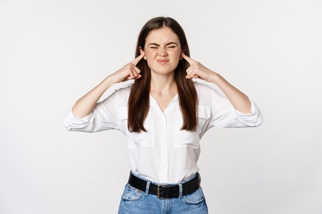 Annoyed girl shut ears from loud sound, discomfort of hearing smth noisy, standing bothered against white background.