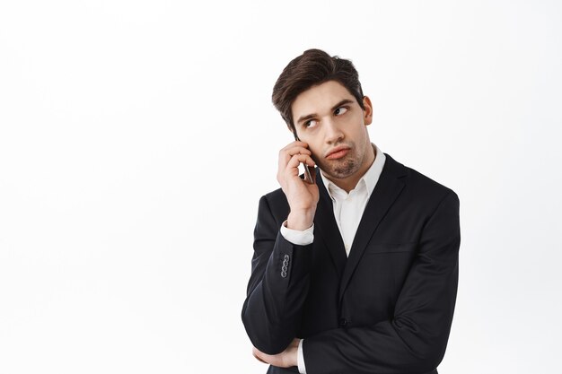 Annoyed guy talking on phone and roll eyes, tired of boring conversation, boring person on call, standing in black suit against white wall