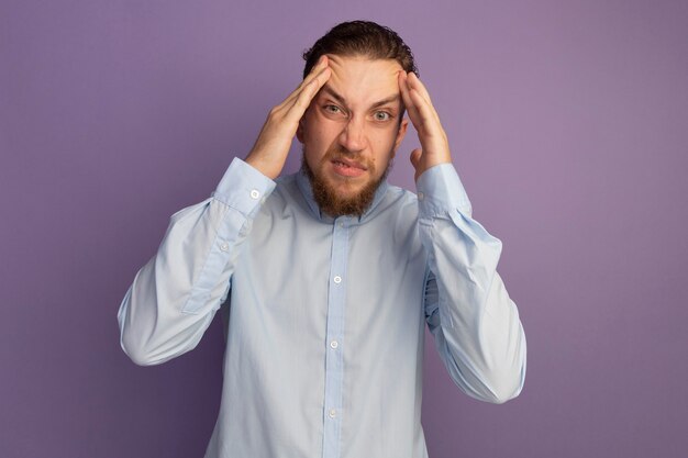 Annoyed handsome blonde man puts hands on forehead isolated on purple wall