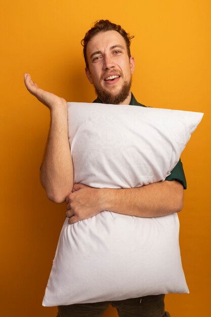 Annoyed handsome blonde man stands with raised hand and holds pillow isolated on orange wall