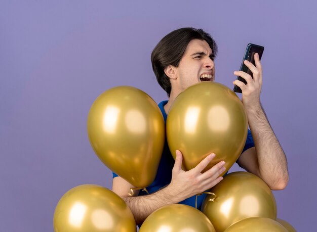 Annoyed handsome caucasian man stands with helium balloons holding and looking at phone 