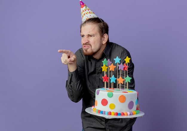 Annoyed handsome man in birthday cap holds cake and points at side isolated on purple wall