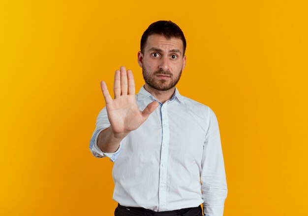 イライラするハンサムな男のジェスチャーは、オレンジ色の壁に分離された手のサインを停止します