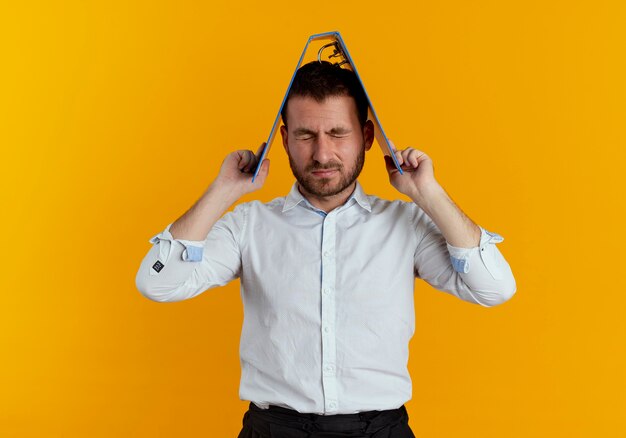 Annoyed handsome man holds file folder over head with closed eyes isolated on orange wall