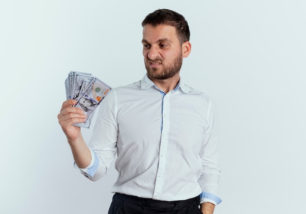 Annoyed handsome man holds and looks at money isolated on white wall