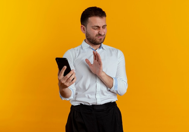 Annoyed handsome man holds phone isolated on orange wall