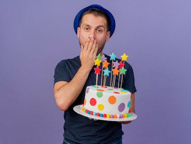 Annoyed handsome man wearing blue hat puts hand on mouth and holds birthday cake isolated on purple wall