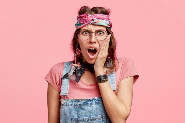 Free photo annoyed hippy female with negative facial expression, keeps hand on cheek, opens mouth widely, being discontent with latest news, poses against pink wall. irritated hippy woman indoor.