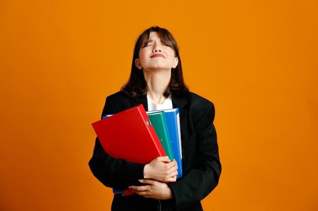 Annoyed holding folders young beautiful female wearing black jacket isolated on orange background