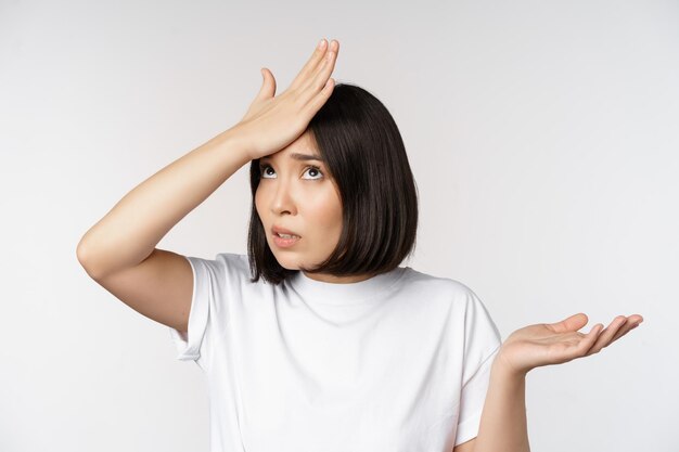 Annoyed korean girl facepalm slap forehead and shrugging confused by smth standing over white background