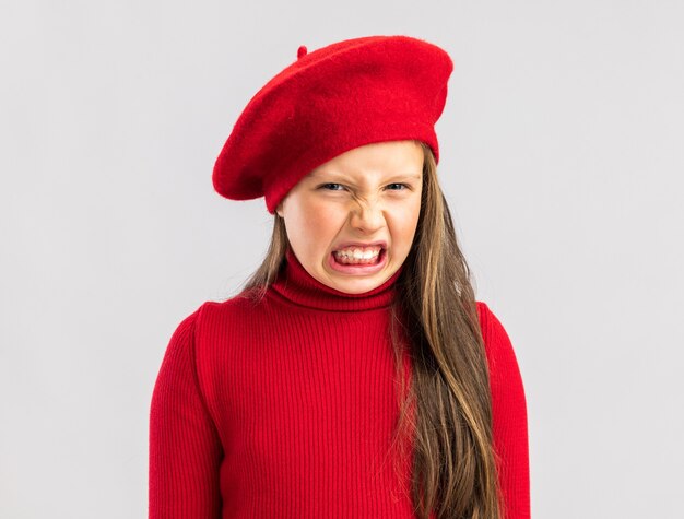 コピースペースで白い壁に分離された赤いベレー帽を身に着けているイライラする小さなブロンドの女の子