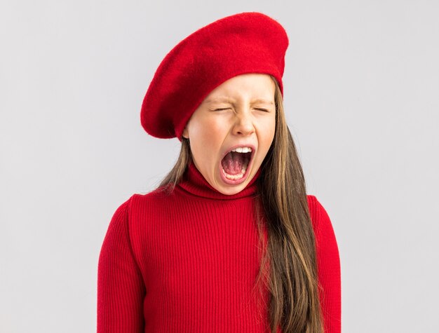 Annoyed little blonde girl wearing red beret keeping eyes closed and screaming isolated on white wall