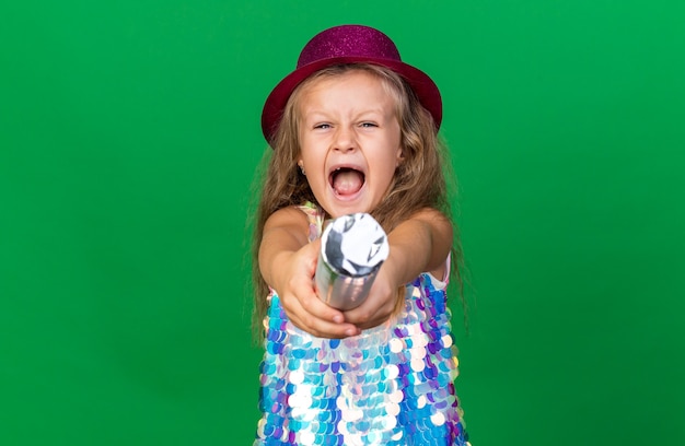 annoyed little blonde girl with purple party hat holding confetti cannon isolated on green wall with copy space