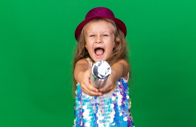 コピースペースで緑の壁に分離された紙吹雪の大砲を保持している紫色のパーティハットでイライラする小さなブロンドの女の子