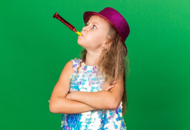 annoyed little blonde girl with purple party hat standing with crossed arms blowing party whistle and looking at side isolated on green wall with copy space