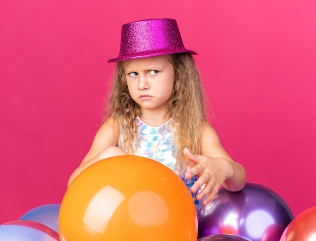 Free photo annoyed little blonde girl with purple party hat standing with helium balloons and looking at side isolated on pink wall with copy space