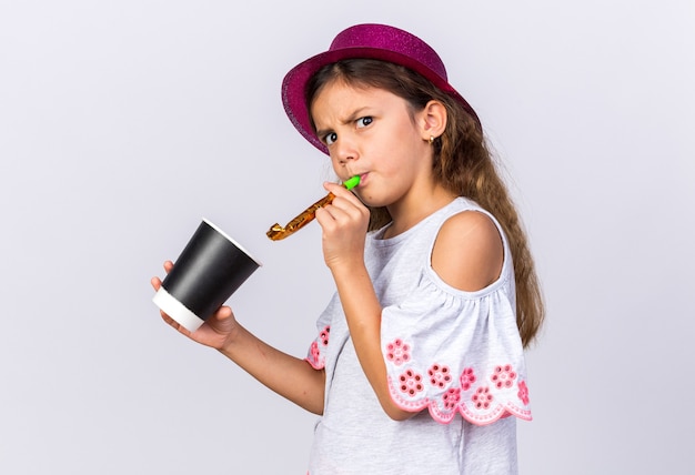 Free photo annoyed little caucasian girl with purple party hat blowing party whistle and holding paper cup isolated on white wall with copy space