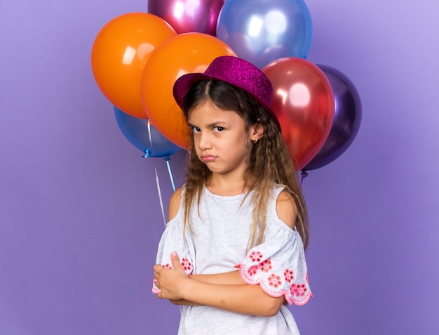 annoyed little caucasian girl with violet party hat standing with crossed arms in front of helium balloons isolated on purple wall with copy space