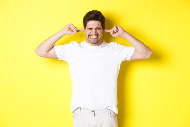 Annoyed man grimacing and shutting ears, complaining on loud noise, standing against yellow background