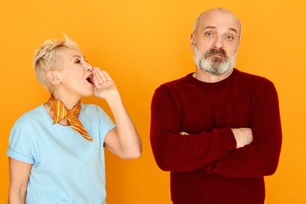 Annoyed mature female with short blonde hair holding hand at her mouth shouting something at husband who is ignoring, not listening to her, keeping arms folded on his chest. Relationships concept