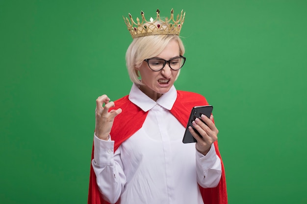眼鏡と王冠を身に着けている赤いマントのイライラした中年の金髪のスーパーヒーローの女性は、コピースペースで緑の壁に隔離された空気中に手を保持している携帯電話を保持し、見ています