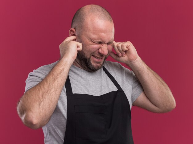 Annoyed middle-aged male barber in uniform closed ears isolated on pink wall