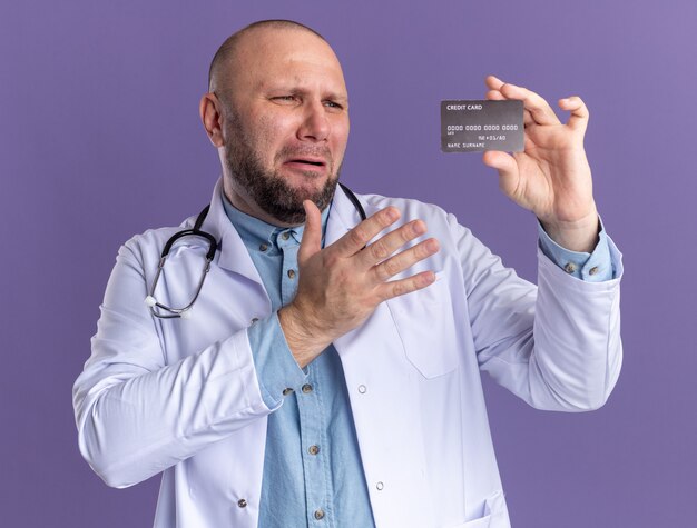 Annoyed middle-aged male doctor wearing medical robe and stethoscope holding and looking at credit card pointing at it 