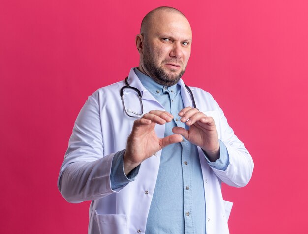 Annoyed middle-aged male doctor wearing medical robe and stethoscope looking at front doing refusal gesture isolated on pink wall