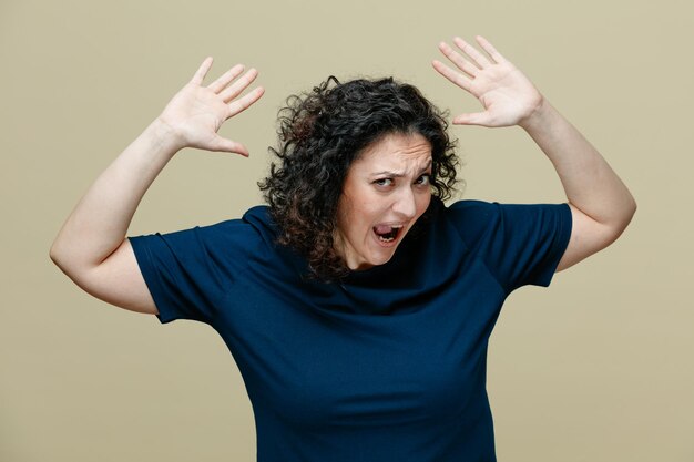 раздраженная женщина средних лет в футболке смотрит в камеру, поднимая руки вверх и крича изолированно на оливково-зеленом фоне