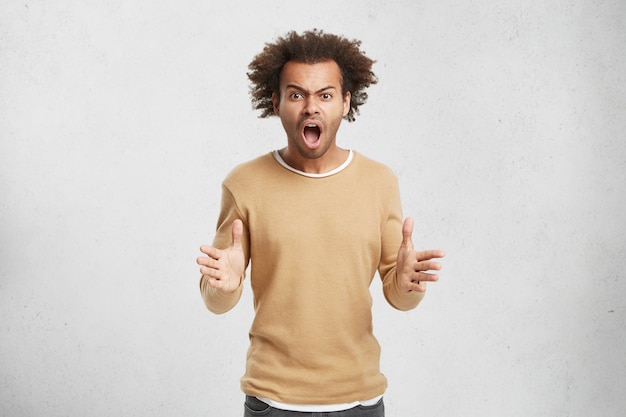 Annoyed mixed race man holds hands in furious gesture, screams loudly as has quarrel