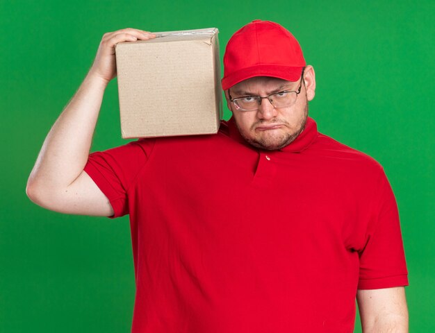 annoyed overweight young deliveryman in optical glasses holding cardboard box on shoulder isolated on green wall with copy space