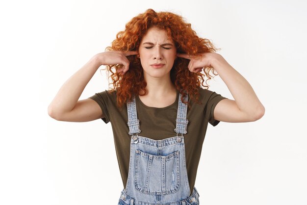 Annoyed and pissed curlyhaired ginger girl plug fingers into ear holes frowning concerned close eyes cringe bothered hear unpleasant loud noise comming from neighbours standing white background