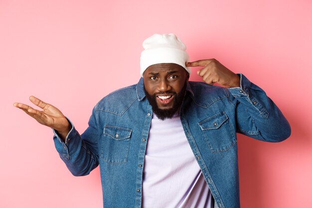 Annoyed and pissed-off african-american man pointing finger at head, scolding someone stupid, staring bothered at camera, pink background