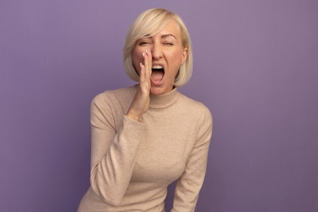 Annoyed pretty blonde slavic woman holds hand close to mouth yelling at someone isolated on purple wall