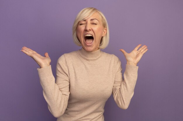 Annoyed pretty blonde slavic woman stands with raised hands screaming with closed eyes on purple