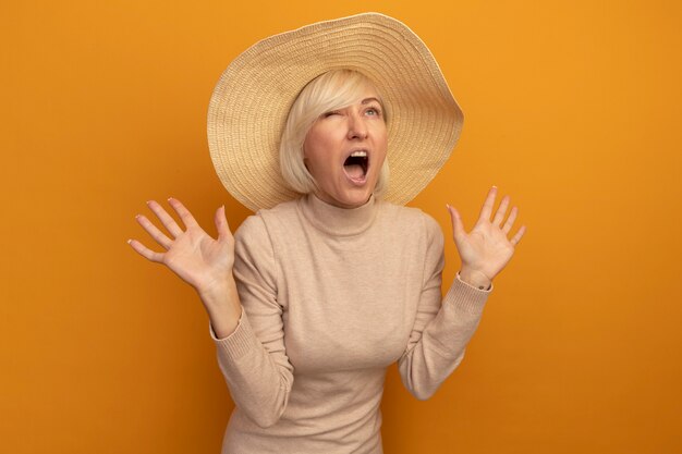 Annoyed pretty blonde slavic woman with beach hat stands with raised hands on orange