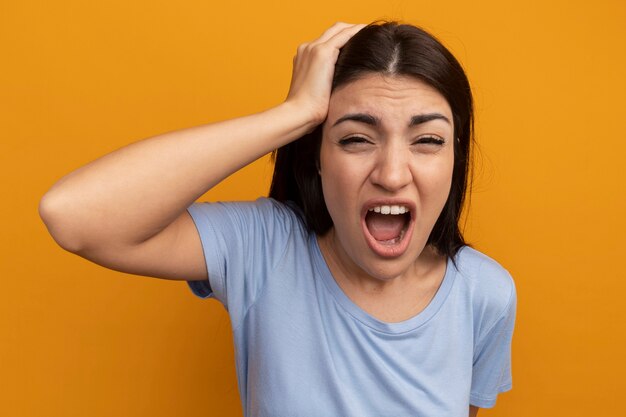 Annoyed pretty brunette caucasian girl puts hand on head and screams on orange