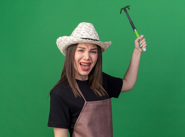 Annoyed pretty caucasian female gardener wearing gardening hat holding hoe rake on green