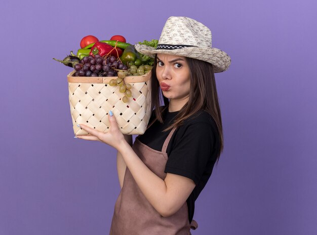 Раздраженная красивая кавказская женщина-садовник в садовой шляпе держит корзину с овощами на плече на фиолетовом