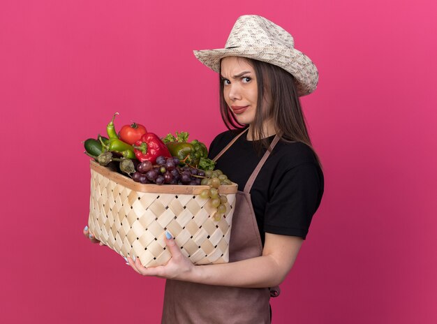 Annoyed pretty caucasian female gardener wearing gardening hat stands sideways holding vegetable basket on pink