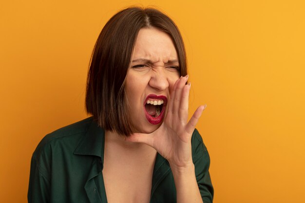 Annoyed pretty caucasian woman holds hand close to mouth pretending to call someone looking at side on orange