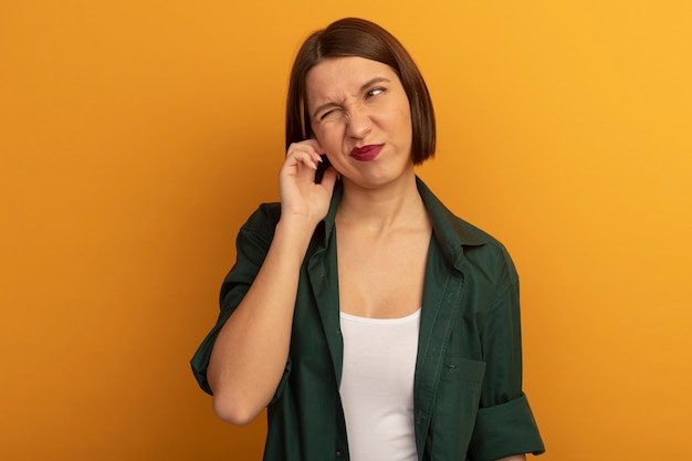Annoyed pretty woman closes ear with finger isolated on orange wall