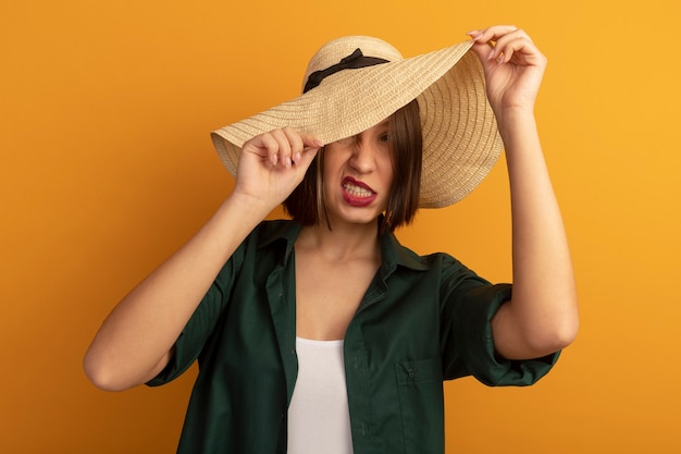 Free photo annoyed pretty woman covers face with beach hat isolated on orange wall