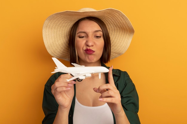 Foto gratuita la donna graziosa infastidita con il cappello della spiaggia tiene ed esamina l'aereo di modello isolato sulla parete arancione