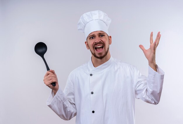 Annoyed professional male chef cook in white uniform and cook hat holding ladle raising hands with aggressive expression feeling irritated standing over white background